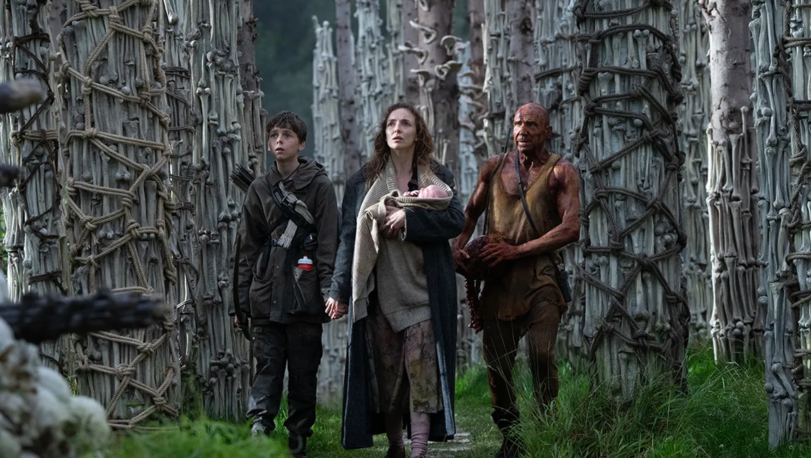 A young boy, a woman, and a shirtless, dirt-covered man stand amidst grass, vines, and trees.