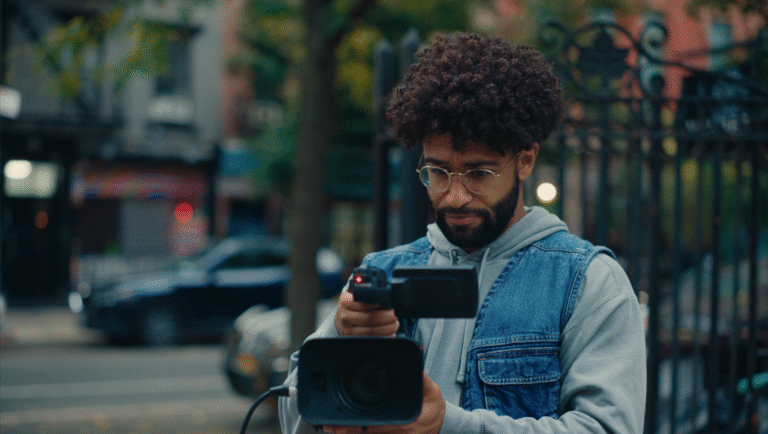 A man with curly hair, glasses, and a denim vest operates a video camera outdoors on a city street, with cars and blurred buildings in the background.