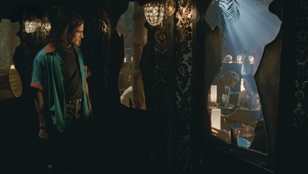 A man in a bright shirt stands in a dimly lit hallway, gazing through uniquely shaped windows at people conversing in a softly illuminated, ornate room as light streams down, creating a dramatic atmosphere.