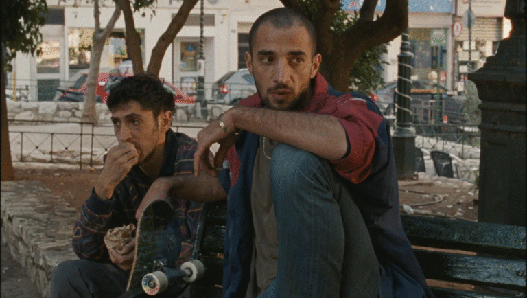 Two men sit on a bench. One eats, the other rests his hand on a skateboard. Urban scene.