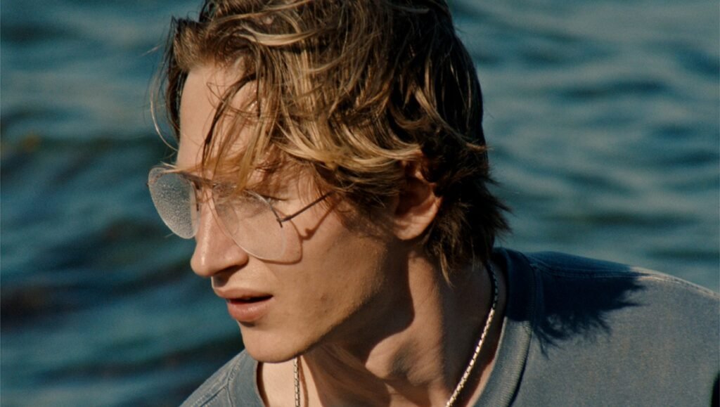 Man with blonde hair, wearing aviator sunglasses, looking out over water. Summer fashion.