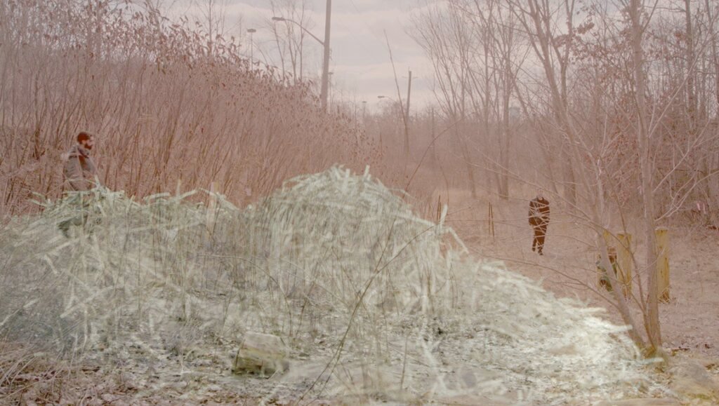 Surreal outdoor scene. Man stands near a pile of hay, another figure is tied to a tree. Gangsterism concept.