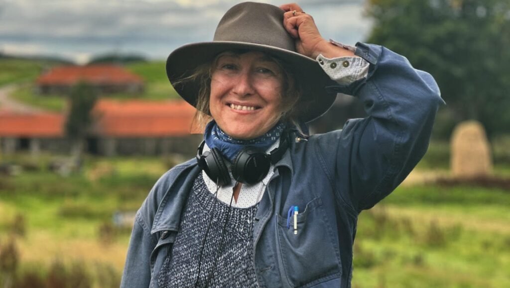 Don’t Break the Spell: An Interview with Athina Rachel Tsangari Athina Rachel Tsangari headshot. Smiling woman with hat, headphones, and blue jacket in a rural landscape. Filmmaker portrait.