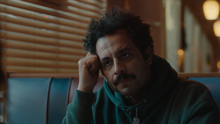 Pensive man with curly hair and mustache, wearing a hoodie. Moody lighting, thoughtful expression.