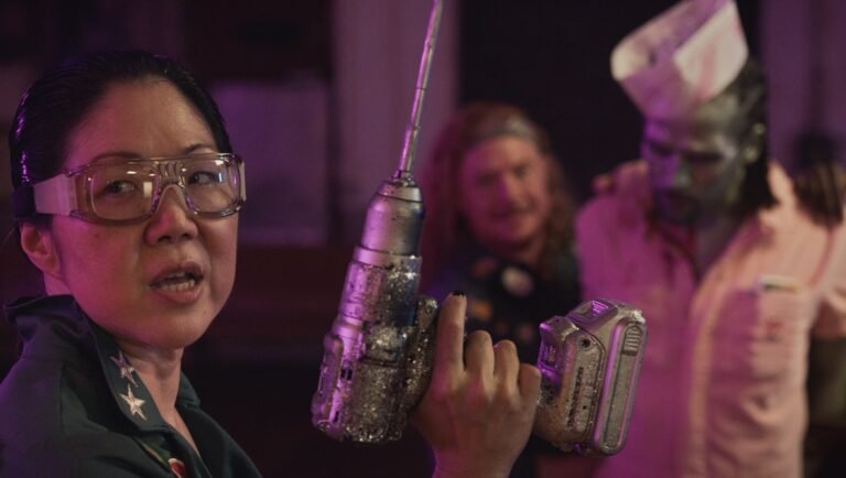 Woman with safety glasses holds drill. Background figures include zombie and person with long hair.
