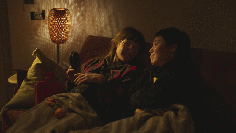 Cozy scene: Two people on a couch, warm lighting, blanket, pillow, hot water bottle. Intimate moment. The Plant from the Canaries film.