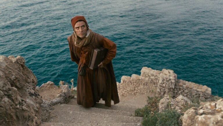 Actor Oscar Isaac in In the Hand of Dante. Man in period costume with book on stone steps by the sea.