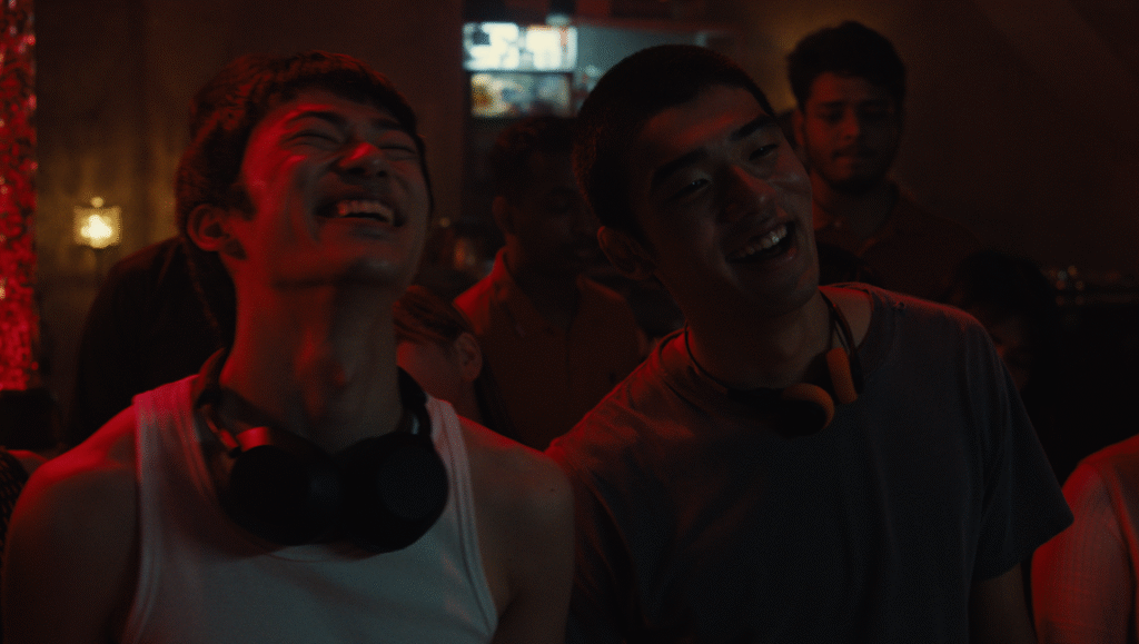 Neo Sora interview image: Two smiling men with headphones around their necks in a dimly lit room.