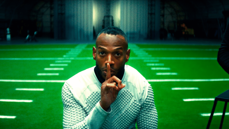Justin Tipping's Him review. Man with finger to lips on a football field. Shhh!