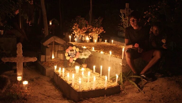 Laguna film still: Night cemetery scene with candles, flowers, crosses, and two people. Venice '25 review.