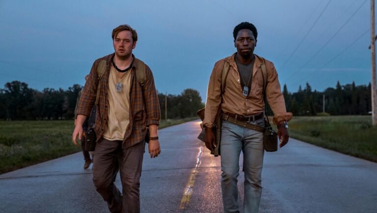 Two men walk along a road. Backpacks, water bottles, and tags are visible. Dusk lighting. Long walk.