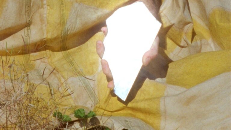 Abstract art: Hand holding white rectangle, yellow fabric, grass. Nature and human element.