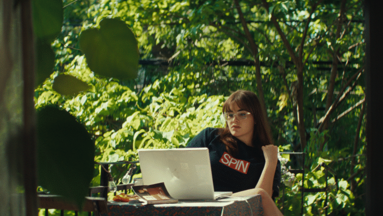 Woman with laptop on balcony. Green trees, natural light. Mile End Kicks.