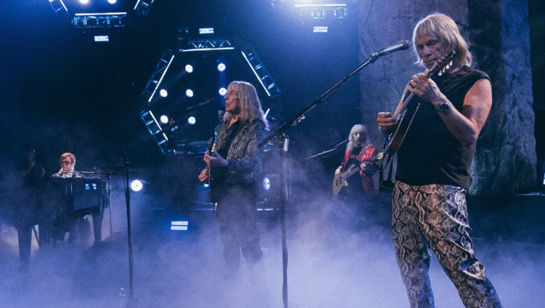 Spinal Tap II still. Band members perform on stage in a smoky blue light. Rock and roll.