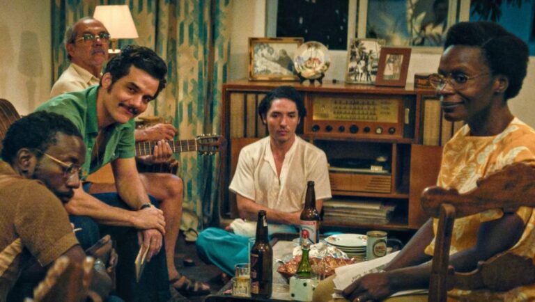 The Secret Agent movie scene. Group of people in a living room with guitar, drinks, and vintage decor. NYFF '25 review.