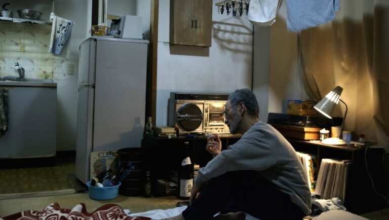 Still from Escape (Masao Adachi, NYFF '25). Elderly man smoking indoors, vintage boombox, cluttered room.