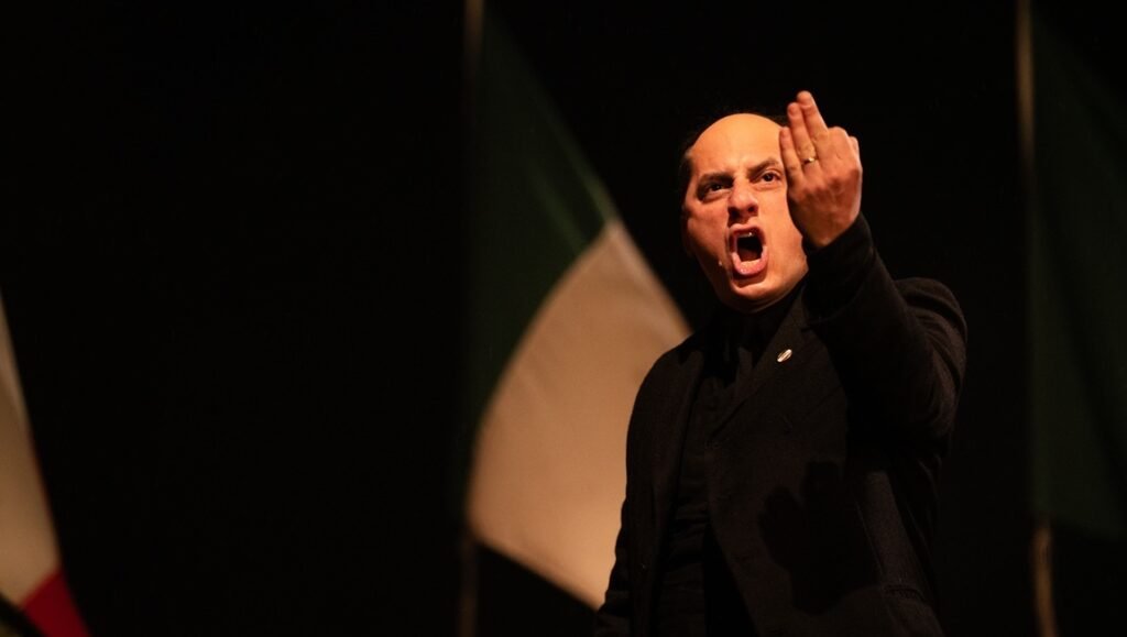 Actor portraying Mussolini in M: Son of the Century. Bald man in black suit gesturing, Italian flag backdrop.