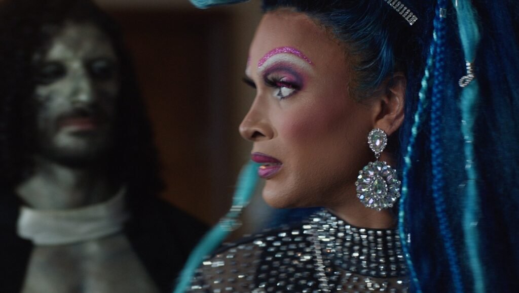 Queens of the Dead still: Tina Romero as a drag queen with blue hair, bold makeup, and sparkling jewelry.
