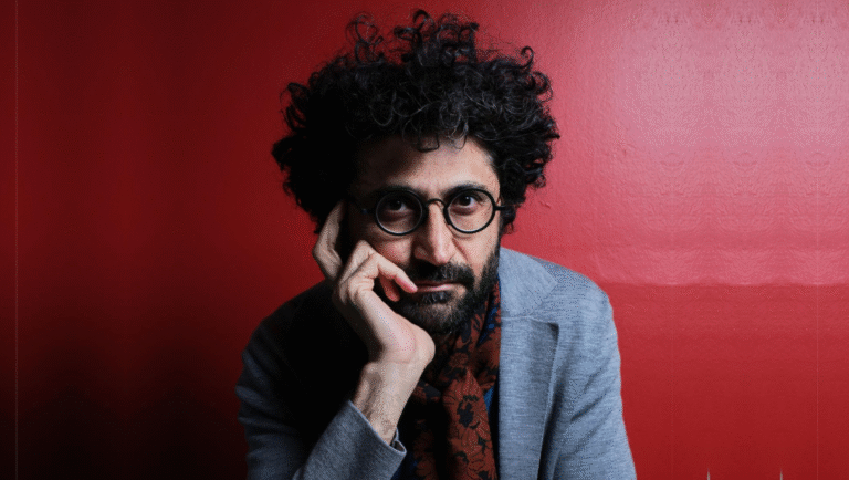 Alireza Khatami portrait. Man with curly hair, glasses, beard, and gray jacket against a red background. Ferda Demir.