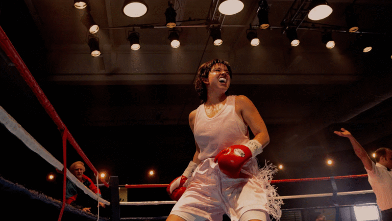 Christy, a boxer, celebrates in the ring. David Michôd film review. Woman with red boxing gloves, white shorts, and a mouthguard.