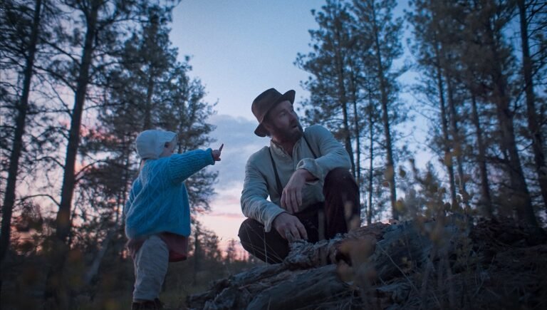 Train Dreams movie scene: Father and child in a forest at dusk. The child points, and the father looks up.