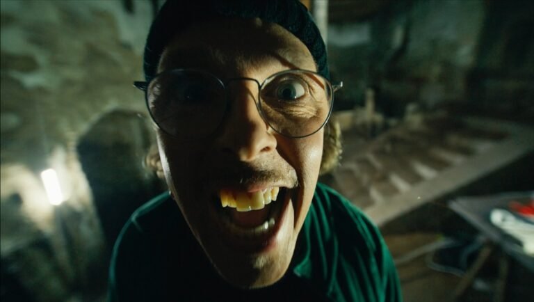 Anniversary film review: Close-up of a man with glasses and a beanie, smiling widely. Jan Komasa film.