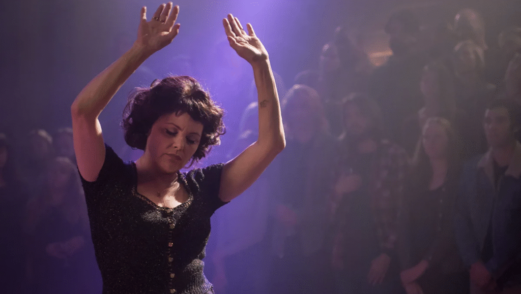 The Return: Woman with raised arms in concert crowd, purple lighting. Dark hair, dark outfit, audience in background.