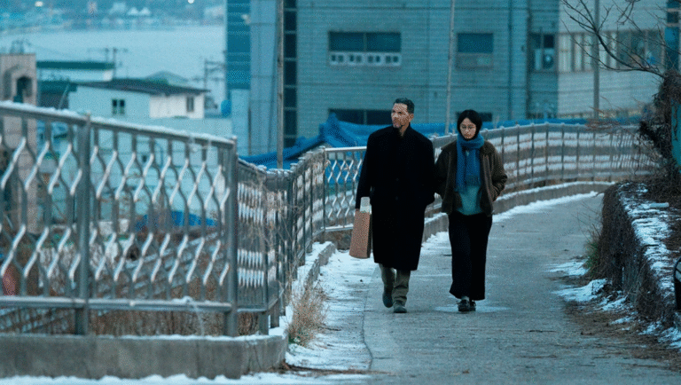 Winter in Sokcho: Man and woman walk on a snowy bridge in Sokcho, South Korea, captured in a still from the Koya Kamura review.