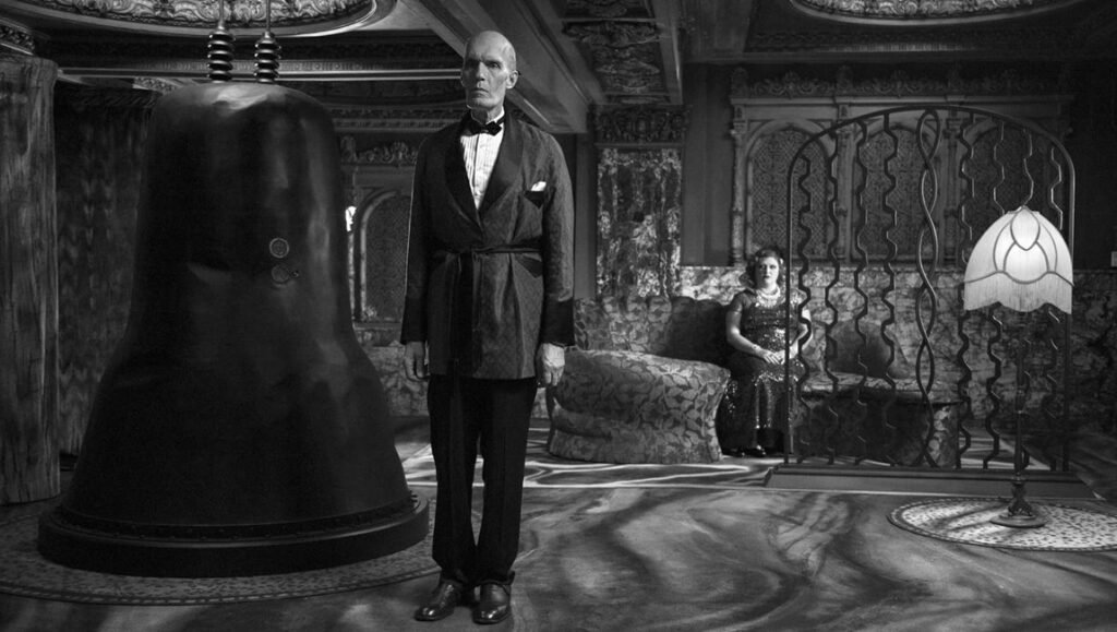 The Return: Black and white photo of a tall man in a suit standing by a large machine, with a woman seated in the background.