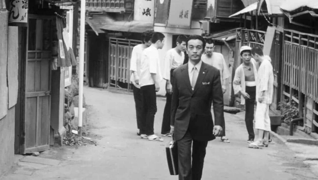 Black and white scene from the movie Flashback, featuring a suited Japanese man walking with a briefcase.