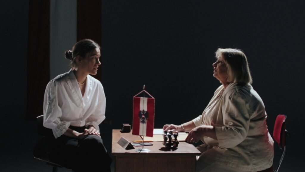 Austrian racism discussion: Two women face each other across a desk with an Austrian flag, highlighting themes of identity.