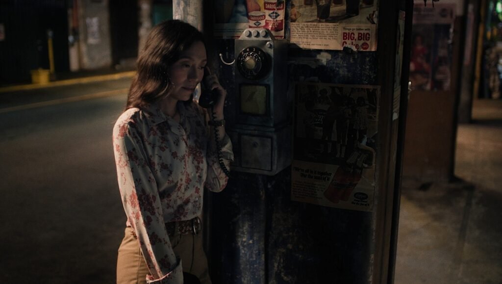 Woman using vintage payphone in urban setting. Nostalgic film still with floral shirt and retro vibe, warm lighting.