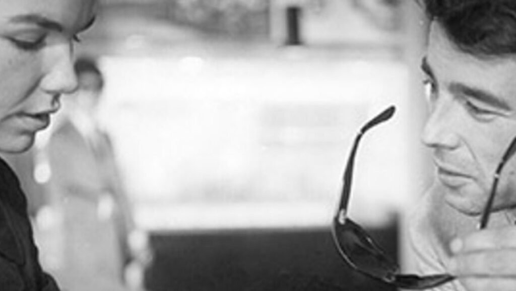 Joë Caligula black and white portrait. Man holding sunglasses, woman in background. Stylish, artistic, classic.