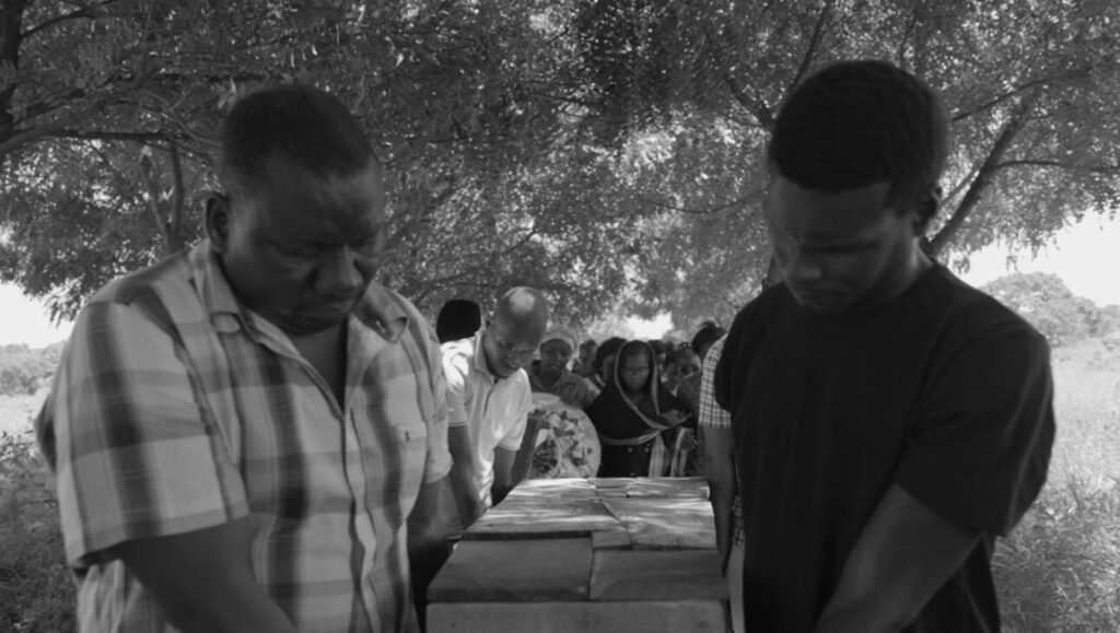 IFFR '26 Review: The Prophet film still. Two men mourn over a casket, with a crowd of mourners in the background.