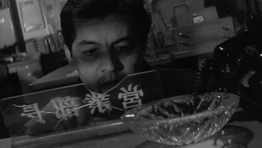 Kinji Fukasaku's 'The Threat' (1966) scene: Post-war salaryman at desk. Japanese film noir, black and white image.