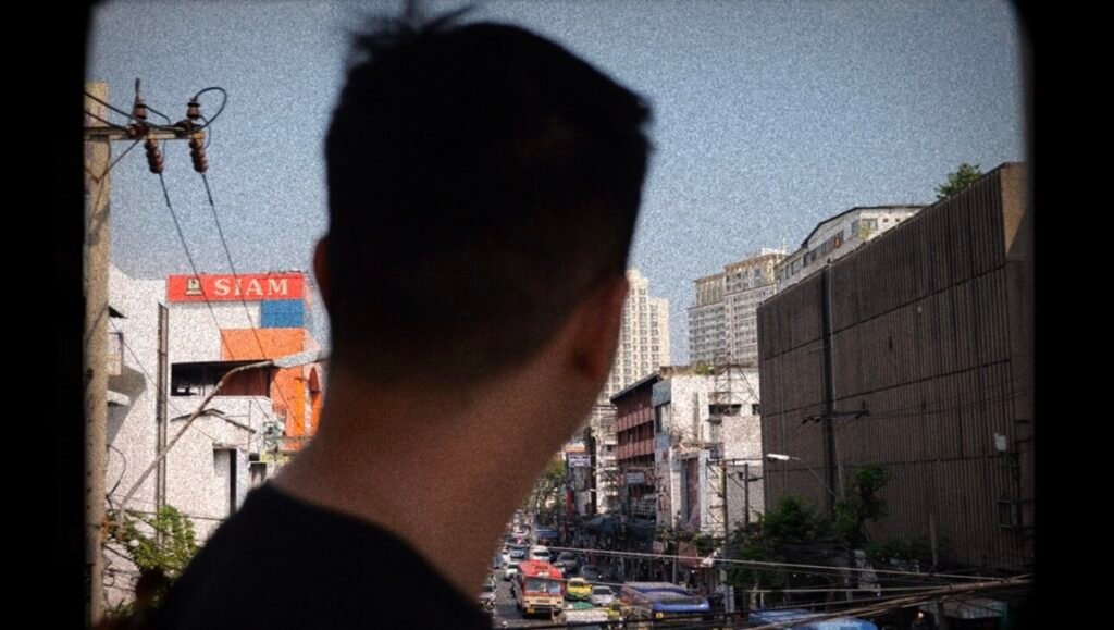 IFFR 2026 film still: Man looks out over urban Bangkok street, featuring 'SIAM' sign, buildings, and street traffic.