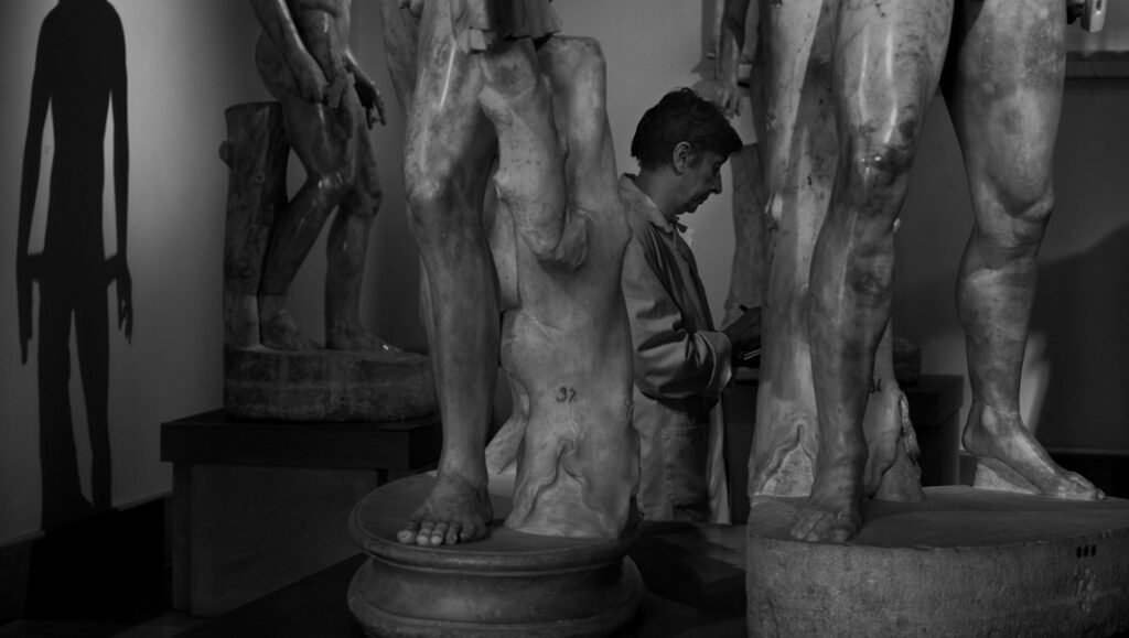 Gianfranco Rosi interview: Man amidst classical sculptures in black and white, examining artistry in detail. "Sotto le Nuvole".