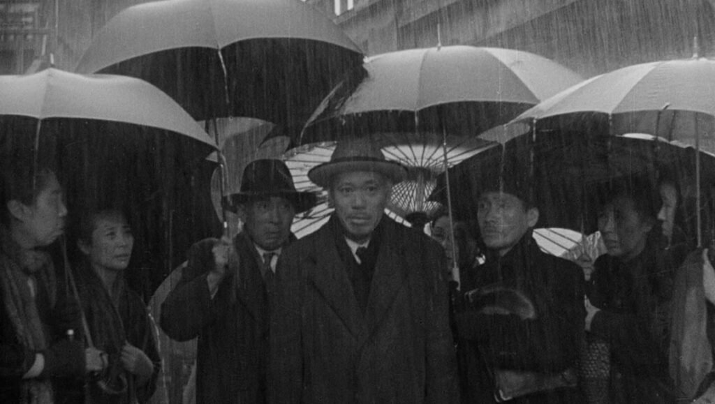 Ikiru film still: Akira Kurosawa movie scene shows people with umbrellas in the rain, classic Japanese cinema.
