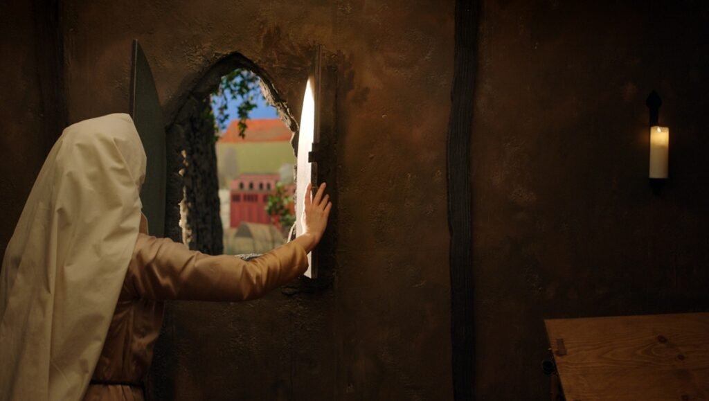 Caroline Golum interview: Woman in historical attire looking out a window at a town, lit by candlelight in a rustic room.