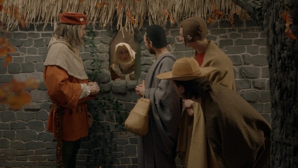 An interview scene: People in medieval costumes gather at a stone wall with a small window, peering inside.
