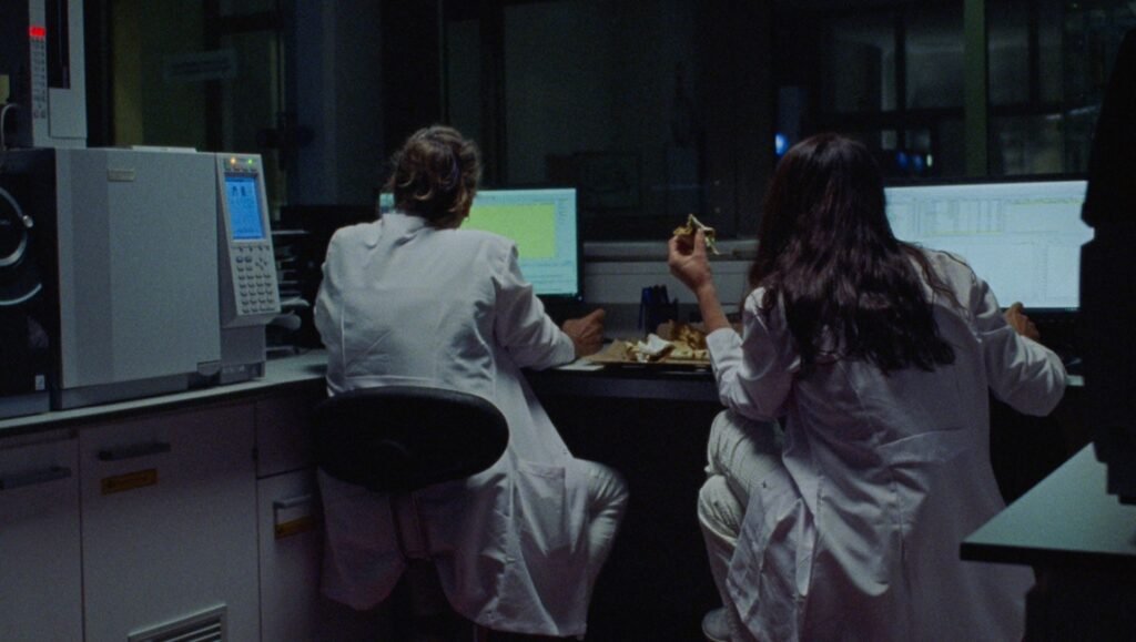 Two weary lab assistants in white coats work at computers, eating during "The Weary Hours of Two Labs Assistants."