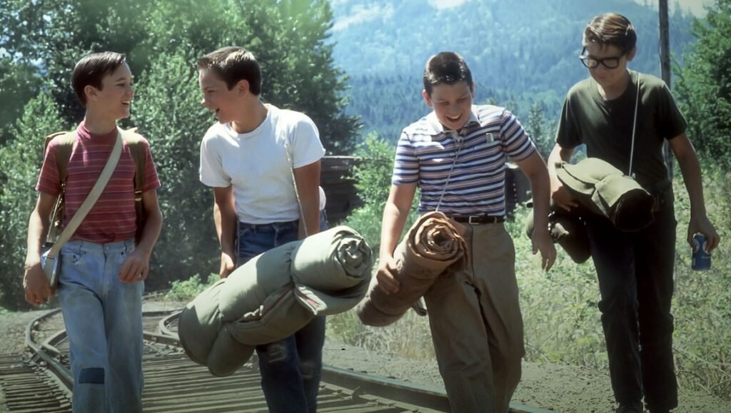 Stand by Me 40th Anniversary: Four boys walk along train tracks carrying camping gear, laughing in a scene from the movie.