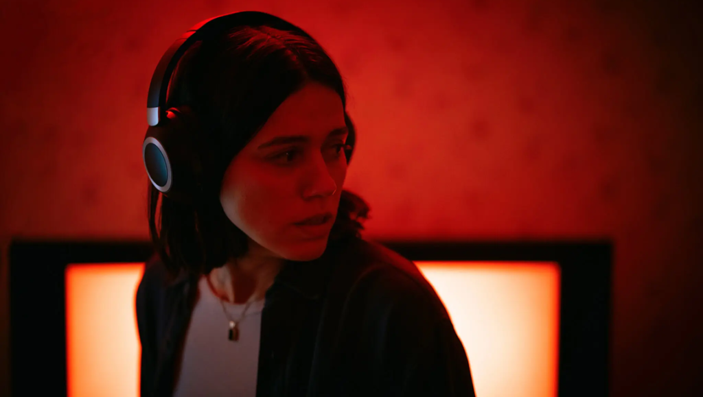 Undertone movie scene: A woman with headphones looks concerned against a red-lit backdrop and a static TV screen.