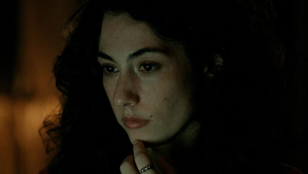 Sophy interview still: Close-up of a woman with dark curly hair thoughtfully gazing downward, finger to chin.