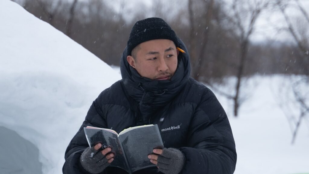 Director Syo Miyake in winter scene holding Montbell notebook, wearing black puffer jacket and beanie with pencil, snowy backdrop