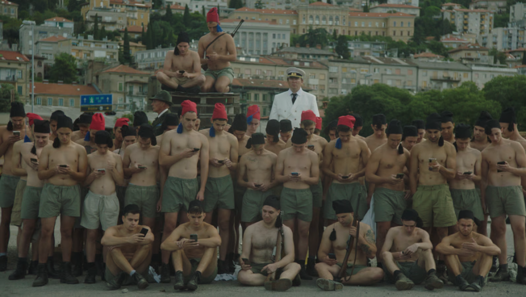 Fiume essay: Group of shirtless men in fezzes using phones, supervised by a man in white uniform; historical reenactment.