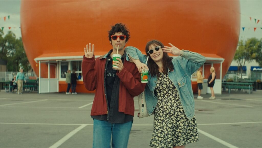 Mile End Kicks film scene: A man and woman pose with orange drinks in front of a giant orange building.