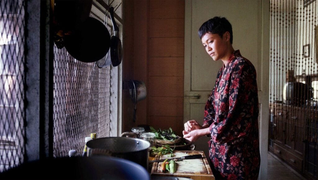 Morte Cucina film still: Woman in floral robe preparing food in Brooklyn kitchen. Indie film scene, Z-grade director's movie.