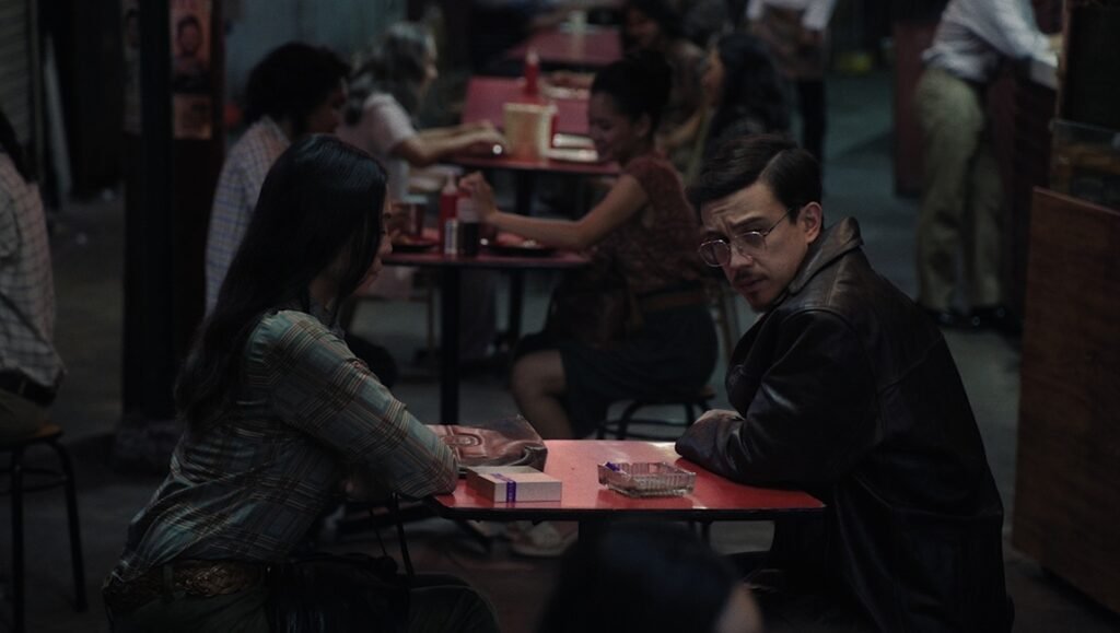 Isabel Sandoval film scene: A man in a leather jacket sits with a woman at a table, cinematic artifice in play.