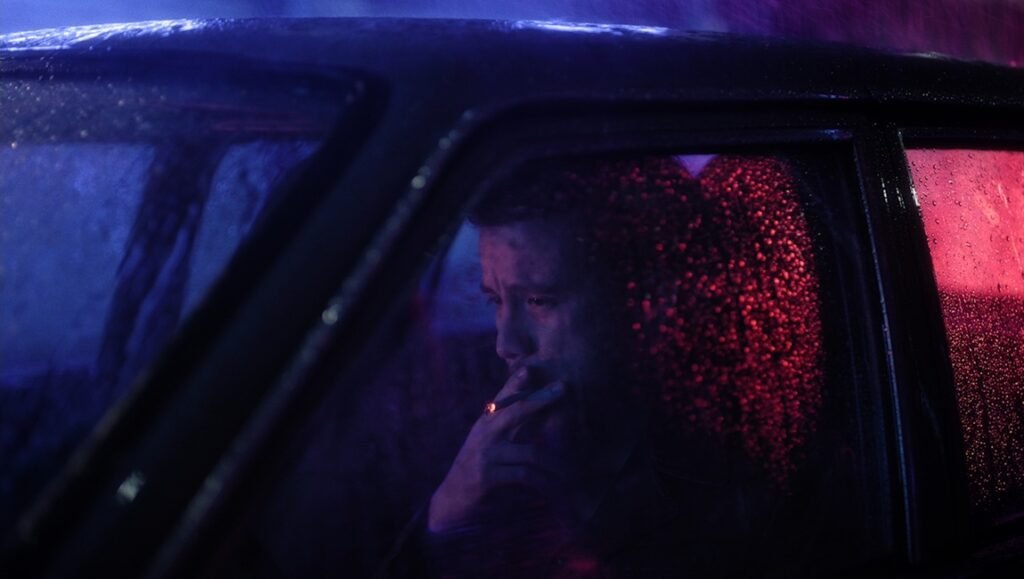 Isabel Sandoval film still: Man smokes in car at night, rain on window, neon lights. Cinema artifice, film noir.